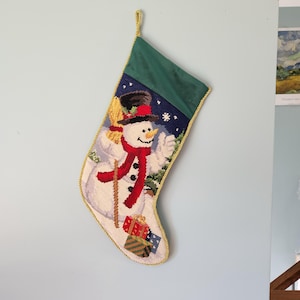 K&ouml;nnte beinhalten: Ein gr&uuml;ner und wei&szlig;er Weihnachtsstrumpf mit einem Schneemann, der einen Besen und Geschenke h&auml;lt. Der Strumpf hat einen goldenen Besatz.