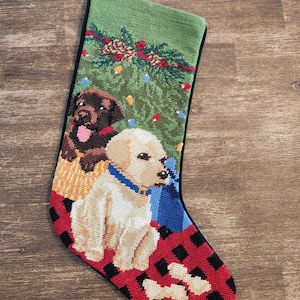 May include: A Christmas stocking with a red and black plaid cuff features two adorable puppies. One puppy is brown and the other is white. The stocking is decorated with a green Christmas tree, red and white ornaments, and pine cones.