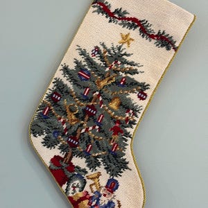 Könnte beinhalten: Ein cremefarbener Weihnachtsstrumpf in Stickoptik mit einem detaillierten grünen Tannenbaum, geschmückt mit Ornamenten, einem goldenen Stern und Girlanden. Unter dem Baum liegen verpackte Geschenke und eine Nussknackerfigur. Goldene Kordel.