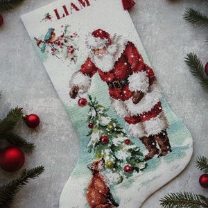 Könnte beinhalten: Ein weißer Weihnachtsstrumpf aus Stoff mit einem Kreuzstichmotiv, das den Weihnachtsmann beim Schmücken eines Tannenbaums zeigt, mit einem kleinen Fuchs an der Basis. Ein blauer Vogel sitzt auf einem Zweig. Der Name LIAM ist oben rot eingestickt.