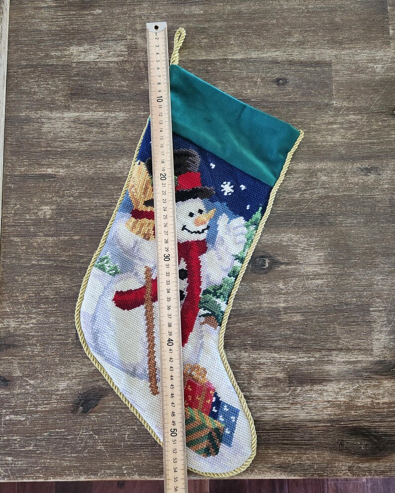 K&ouml;nnte beinhalten: Ein wei&szlig;er Weihnachtssocken mit einem Schneemann-Motiv. Der Socken ist mit goldener Litze verziert und hat einen gr&uuml;nen Samt-Manschetten. Der Schneemann tr&auml;gt einen roten Schal und Hut und h&auml;lt ein Geschenk. Der Socken ist 142 cm lang.