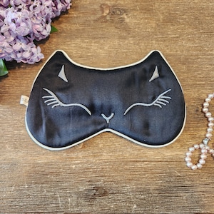 May include: Black sleep mask with a white cat face embroidered on it. The mask is on a wooden surface with a pearl necklace next to it.