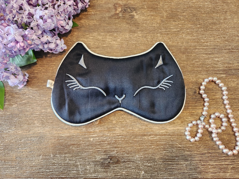 May include: Black sleep mask with a white cat face embroidered on it. The mask is on a wooden surface with a pearl necklace next to it.