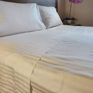 Puede incluir: Sábanas de cama blancas a rayas con dos almohadas blancas sobre un marco de cama de madera.