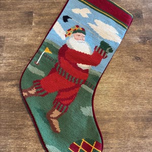 May include: A needlepoint Christmas stocking featuring a depiction of Santa Claus playing golf on a green course. Santa wears a red sweater, red knickers, and a plaid hat. The background shows a blue sky with white clouds, a yellow flag, and a golf club.