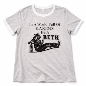 May include: A light gray t-shirt with a white collar and sleeve trim. The shirt features a black graphic of a woman in a cowboy hat with a bottle and drink, and the text "In A World Full Of Karens Be A Beth."