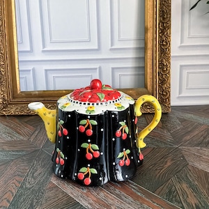 Op de afbeelding: Een keramische theepot met een zwarte basis, een wit stippenpatroon en rode kersenmotieven. Het deksel is gevormd als een tros rode kersen. De theepot heeft een geel handvat, een gele tuit met kleine zwarte markeringen en een geschulpte rand.
