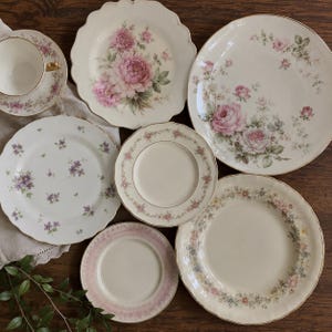 Puede incluir: Una colección de seis platos de cerámica y una taza de té con platillo, con varios patrones florales en tonos rosa, morado y verde. Las piezas tienen bordes festoneados o lisos, algunas con borde dorado, dispuestas sobre una superficie de madera.