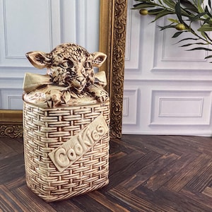 May include: A ceramic cookie jar featuring a sculpted lamb head resting on a lid, with a woven basket texture on the body. The word Cookies is embossed diagonally across the front in raised lettering. The piece has a cream glaze with brown accents.