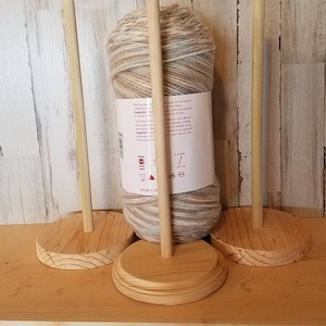 May include: Three wooden yarn holders with a natural finish. Each holder has a round base and a vertical dowel. A ball of variegated yarn in shades of beige, brown, and gray is displayed on one of the holders. The yarn label is visible.