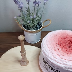 May include: A wooden yarn swift with a light-colored base and a spool-like center, next to a ball of pink and white ombre yarn. A small white bucket with lavender flowers sits in the background.