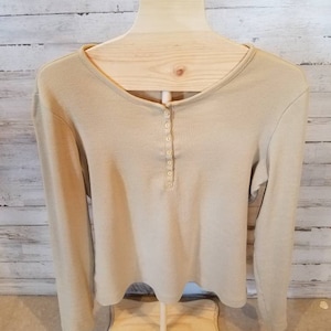 May include: A long-sleeved, beige shirt with a scoop neck and a button-up placket. The shirt is displayed on a wooden mannequin against a light-colored, wood-paneled background. The shirt has a ribbed texture.