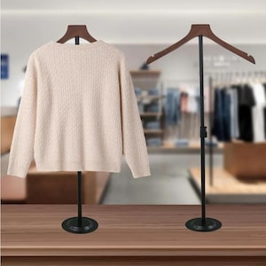 May include: A cream-colored sweater displayed on a black metal clothing rack with a wooden hanger. The sweater has a textured cable knit pattern. Another rack with a wooden hanger stands beside it. The racks are on a wooden surface.