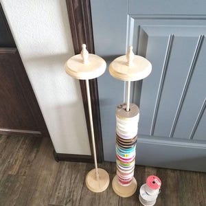 May include: Two wooden ribbon organizers with a natural finish. One holds spools of colorful ribbon, the other is empty. A small stack of ribbon spools sits nearby. The organizers are on a wooden floor.