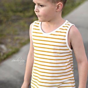 May include: A young boy wearing a white and yellow striped tank top with a blue pair of shorts. The tank top has a round neckline and thin straps.