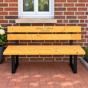 Op de afbeelding: Een houten bank met een natuurlijke lichtbruine afwerking en een zwart metalen frame. De bovenste rugleuning is voorzien van de gegraveerde tekst "Marlene ♡ Marcel" boven de datum "19.06.2026" in een donker scriptlettertype. De bank heeft drie houten latten.