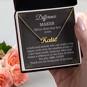 Puede incluir: Un collar dorado con el nombre "Katie" en escritura cursiva, presentado en una caja de regalo negra. La caja presenta las palabras "Difference Maker" y una definición. Se ven rosas rosas en el fondo.