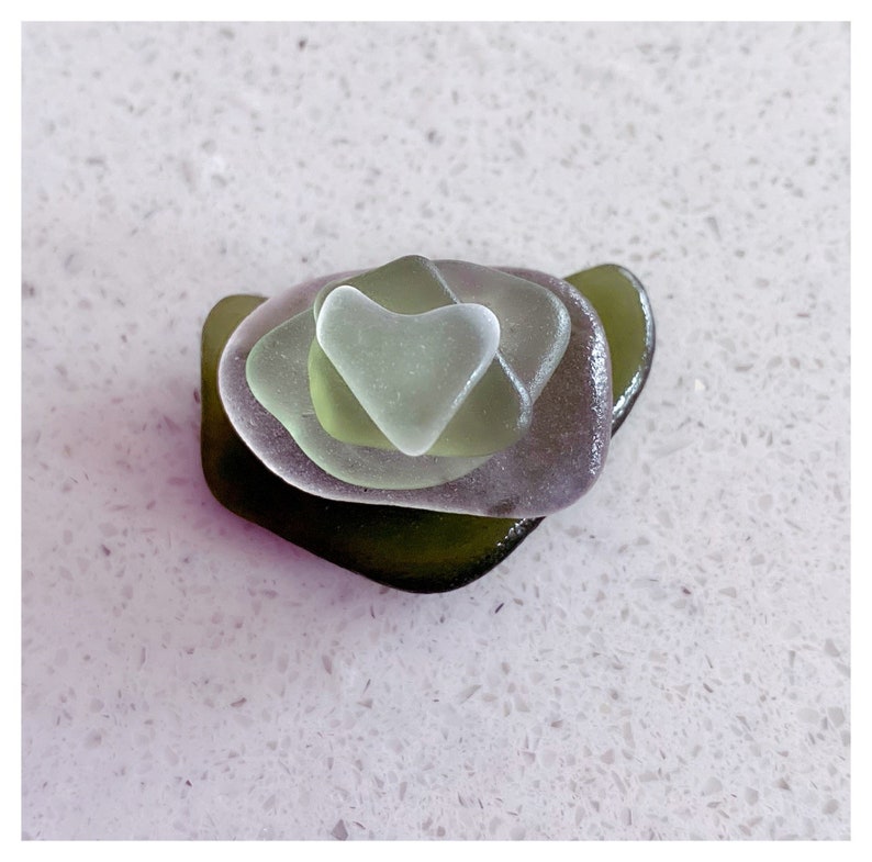 May include: A stack of sea glass in shades of green, with a heart-shaped piece of sea glass on top.