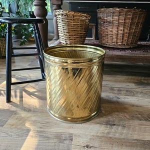 May include: A cylindrical brass waste bin featuring a diagonal ribbed texture across its exterior surface. The metal container has a polished finish with a wide circular opening at the top and a flat base, standing upright as a standalone object.
