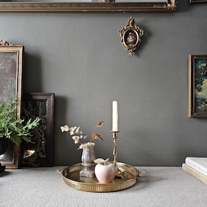 May include: A gold metal tray with a decorative edge holds a pink apple, a small white alabaster vase, and a gold candle holder with a white candle. The tray is on a white surface in front of a grey wall with framed artwork.