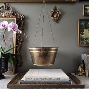 May include: A hanging brass planter with a tapered, cylindrical body and a smooth, metallic finish. The planter is suspended by three thin, interconnected brass chains that meet at a single point above, creating a balanced, open-top vessel.