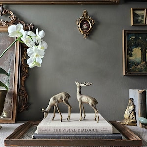 May include: Two brass deer figurines stand on a stack of two books titled The Visual Dialogue and Masters of Painting. One deer is positioned with its head lowered towards the book surface, while the other stands upright with its head tilted upwards.