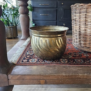 May include: A vintage brass planter with a swirling design. The planter has a wide, fluted rim and sits on a wooden surface. A woven basket and a patterned rug are also visible in the background, suggesting a rustic or antique theme.