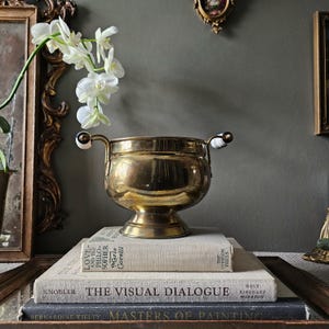 Peut inclure: Un bol en laiton poli avec un corps arrondi et une base sur pied, doté de deux poignées latérales avec des accents en céramique rayés blancs et noirs. Le bol est posé sur une pile de trois livres reliés avec du texte visible sur les tranches.