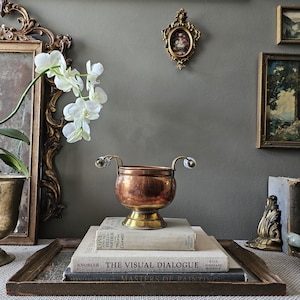 Peut inclure: Un bol en cuivre avec une base en laiton et deux anses incurvées ornées d'accents en porcelaine bleue et blanche. Le bol est posé sur une pile de trois livres à couverture rigide, dont The Visual Dialogue et Masters of Painting.