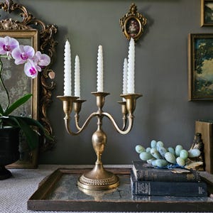 May include: A brass candelabra with five arms holding white spiral taper candles. The metal base features a circular pedestal with decorative ridged detailing. The candelabra stands on a rectangular mirrored tray alongside two stacked books.