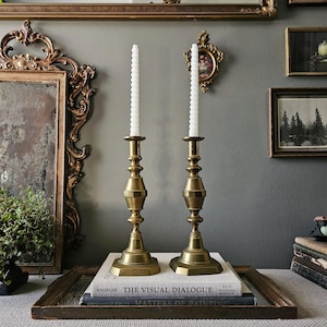 May include: Two brass candlesticks, each holding a white spiral candle, are displayed on a stack of books. The candlesticks have a detailed design and are placed on a wooden tray. The background includes framed artwork and a decorative mirror.