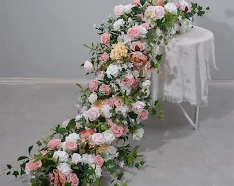 Puede incluir: Una larga guirnalda de flores artificiales en tonos rosa, blanco y crema. La guirnalda está drapeada sobre una mesa blanca y presenta rosas, peonías y otras flores.