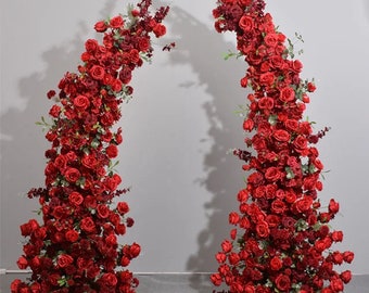 May include: Two tall, red artificial flower arrangements, each made of red roses and greenery, are arranged in a semi-circle shape. The arrangements are on white stands and are against a white wall.