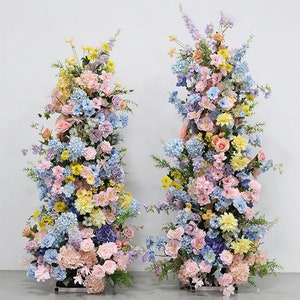 May include: Two tall floral arrangements featuring a mix of pastel-coloured flowers, including pink, blue, yellow, and purple. The flowers are arranged in a cascading style, creating a romantic and elegant look.