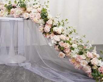 Puede incluir: Una larga guirlanda de flores artificiales en tonos rosa y blanco, con vegetación, que cae en cascada sobre una mesa blanca cubierta de tul. La guirlanda es perfecta para una boda u otro evento especial.