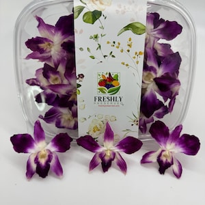May include: A clear plastic container filled with purple and white orchids. Three orchids are placed in front of the container. The container has a label with floral designs and the text "FRESHLY PRESERVED".