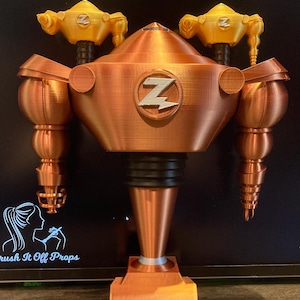 以下が含まれることがあります： 胸にZのロゴが付いた銅色のロボット。ロボットは、各腕にノズルが付いた2本の黄色い腕を持っている。ロボットは黒い表面の上に立っている。Brush It Off Props.