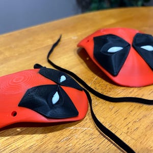 May include: Two red 3D printed Deadpool masks with black eye holes and white eye details. The masks are attached to black cords.
