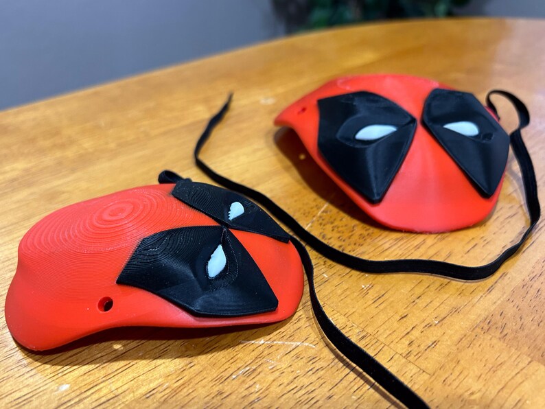 May include: Two red 3D printed Deadpool masks with black eye holes and white eye details. The masks are attached to black cords.