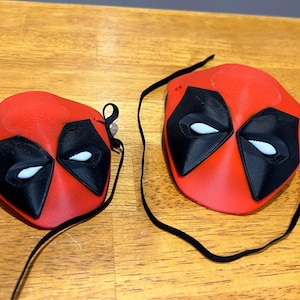 May include: Two red and black Deadpool masks with white eyes. The masks are attached to black elastic straps.