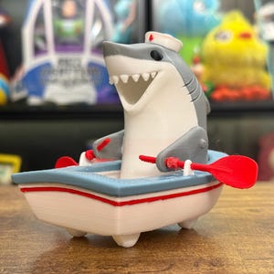 May include: A novelty figurine of a shark wearing a nurse's hat, in a small boat with red oars. The boat is white with a blue rim and red accents. The shark is grey and white, with a friendly expression.