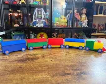 Op de afbeelding: Een meerkleurige speelgoedtrein bestaande uit een locomotief en vier gekoppelde wagons. De locomotief is groen en geel met een roze schoorsteen. De wagons zijn geel, rood, groen en blauw, allemaal met kleine blauwe wielen met roze kernen.
