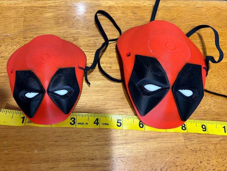 May include: Two red and black Deadpool masks with white eyes. The larger mask has a string attached to it. The masks are on a wooden surface with a measuring tape.