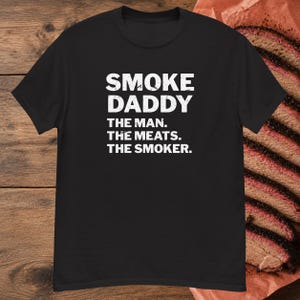 Pu&ograve; includere: Una t-shirt nera a maniche corte con testo bianco in stile vintage che recita SMOKE DADDY THE MAN. THE MEATS. THE SMOKER. La maglietta &egrave; presentata su uno sfondo di fette di carne affumicata adagiate su carta da macellaio marrone.