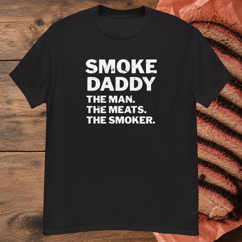 Pu&ograve; includere: Una t-shirt nera a maniche corte con testo bianco in stile vintage che recita SMOKE DADDY THE MAN. THE MEATS. THE SMOKER. La maglietta &egrave; presentata su uno sfondo di fette di carne affumicata adagiate su carta da macellaio marrone.