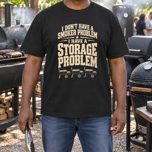May include: A black short-sleeved t-shirt featuring the beige text I DON'T HAVE A SMOKER PROBLEM I HAVE A STORAGE PROBLEM and a graphic of three horizontal barrel-style smokers with wheels centred on the chest.