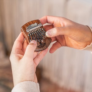 Può includere: Un piccolo kalimba di legno con un design ad albero della vita. Il kalimba ha 10 linguette metalliche ed è tenuto nelle mani di una persona.