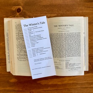 May include: A black and white printed card with the title "The Winter's Tale" and a list of characters from the play. The card is sitting on top of an open book with the same title.