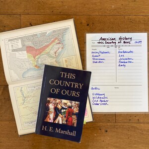 Pu&ograve; includere: Un libro rilegato in blu intitolato "This Country of Ours" di H.E. Marshall. La copertina del libro presenta un gruppo di persone in abiti d'epoca. Sullo sfondo &egrave; visibile un elenco manoscritto di battaglie, persone e luoghi legati alla storia americana.
