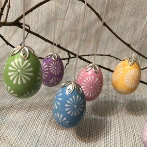 May include: A selection of Easter egg decorations suspended from twigs. The eggs are painted in pastel colours including green, purple, pink, yellow, and blue, each with a white floral pattern. Silver-tone caps and white strings add to the festive appearance.